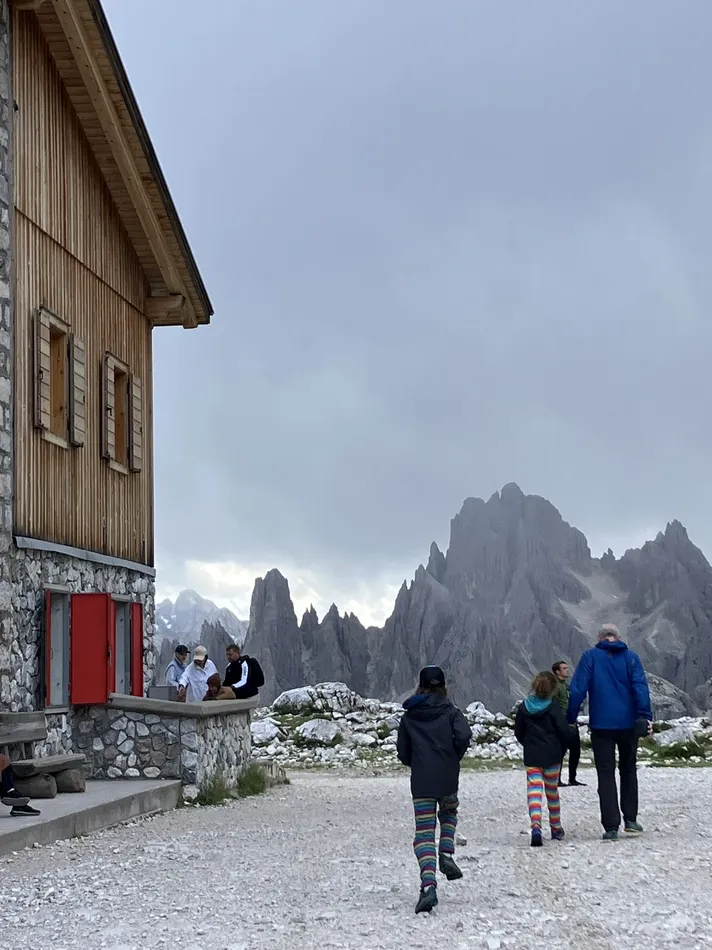 A close-up view of a mountain hut with a wooden facade and red shutters, situated on a rocky pathway. People are gathered outside, with a dramatic backdrop of towering, craggy peaks partially obscured by misty clouds.