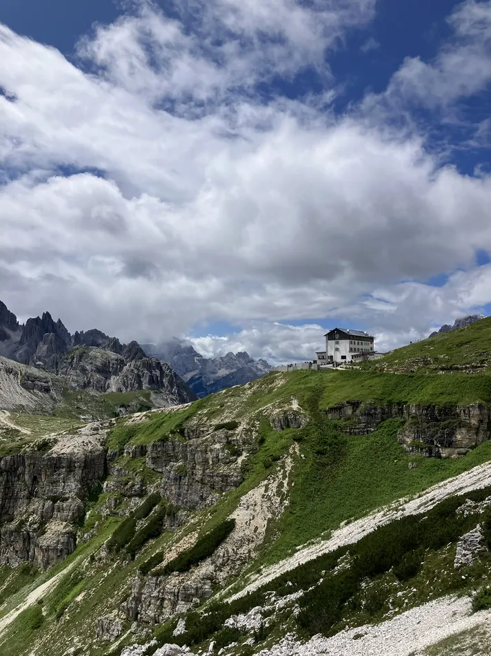 A mountain lodge perched on a grassy ridge surrounded by rugged cliffs, with distant peaks rising under a dramatic sky filled with thick, white clouds. The scene highlights the serene beauty and isolation of the alpine environment.