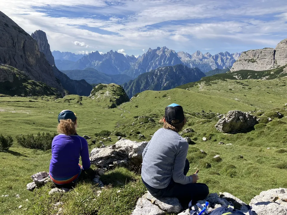 Two kids hut to hut hiking in the Dolomites sit on a grassy hill overlooking a vast, mountainous landscape. The sky is partly cloudy, and the scene includes rugged peaks and lush, green valleys. They are dressed casually, with hats and jackets.