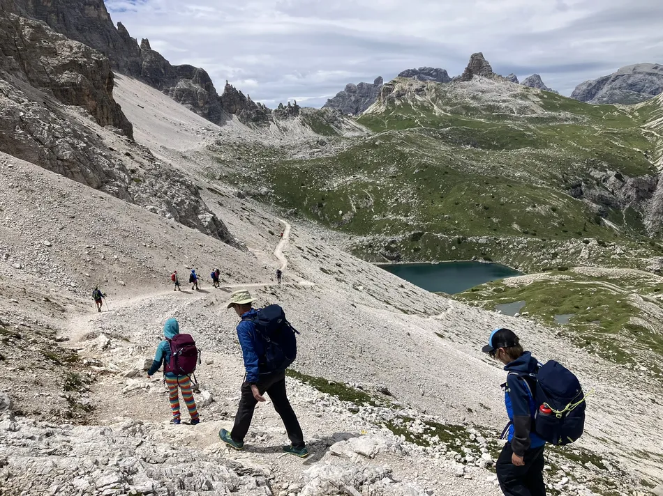 A family hiking along a rocky trail in the Dolomites, with a shimmering alpine lake and jagged peaks in the background. This photo highlights the adventurous spirit of a hut hike in the Dolomites with kids, surrounded by breath taking landscapes.