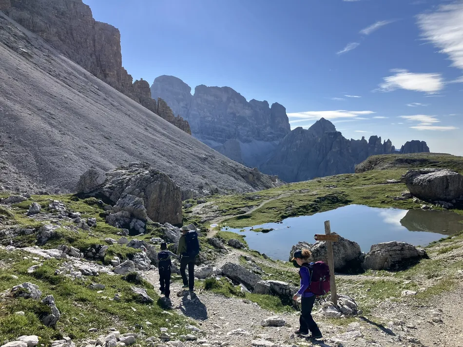 Two hikers walk along a rocky trail near a small reflective pond surrounded by grass and boulders, with jagged mountain peaks rising dramatically in the distance. A wooden trail sign stands by the path, and the bright blue sky enhances the scenic alpine setting.