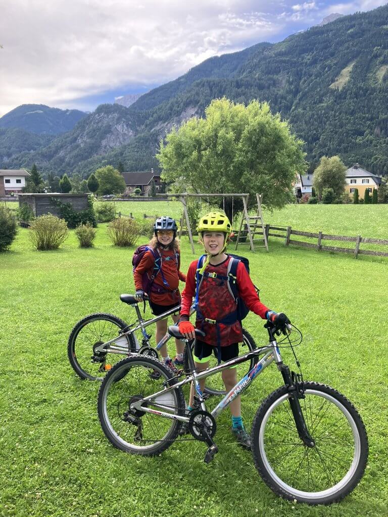 Two children in colorful cycling gear, wearing helmets and backpacks, stand with their bikes on a vibrant green field. Behind them is a wooden swing set, trees, houses, and a backdrop of lush green mountains under a partly cloudy sky.