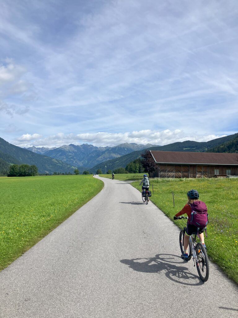 Two cyclists riding on a paved path through a scenic valley with green fields, a wooden barn, and mountains in the distance. The sky is partly cloudy, and the surrounding landscape is lush and vibrant, suggesting a peaceful, rural setting