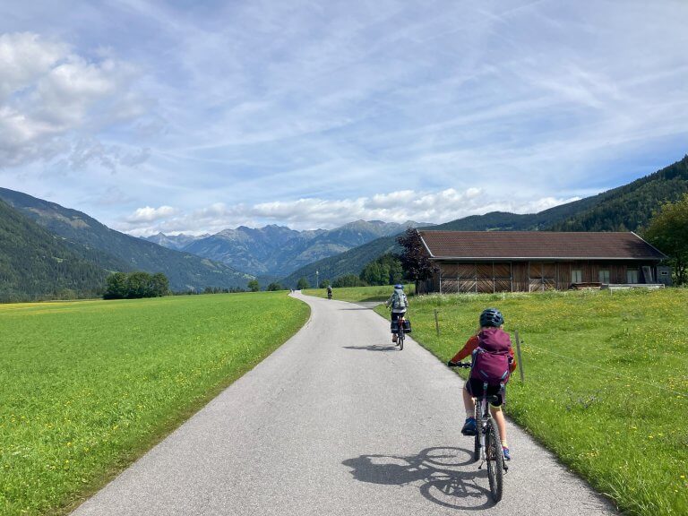 Two cyclists riding on a paved path through a scenic valley with green fields, a wooden barn, and mountains in the distance. The sky is partly cloudy, and the surrounding landscape is lush and vibrant, suggesting a peaceful, rural setting