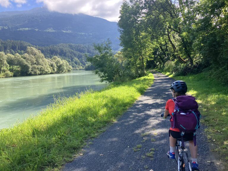 A child wearing a helmet and backpack rides a bicycle on a tree-lined path beside a river, with mountains in the background under a partly cloudy sky.