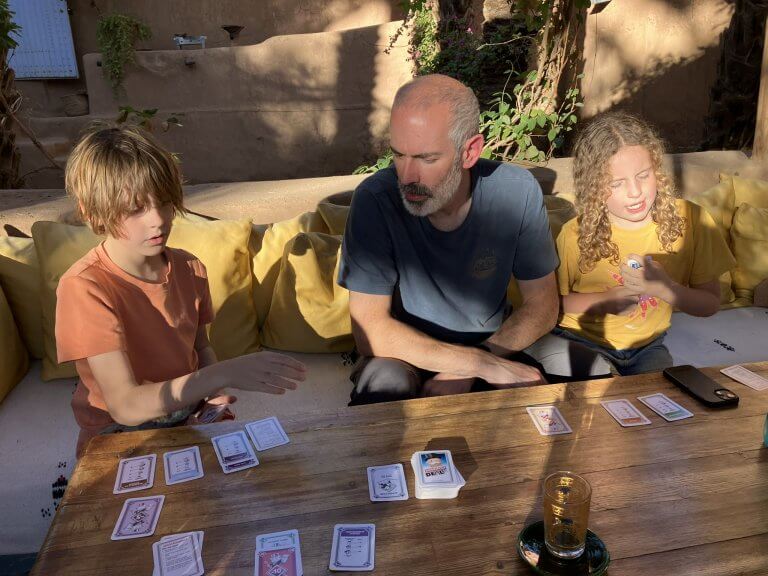 A young boy in an orange shirt, a bearded man in a blue shirt, and a girl with curly hair wearing a yellow shirt are sitting around a wooden table playing a card game. The sunlight casts strong shadows across the table, which has cards laid out, and the trio looks focused on their game. Cushioned seating with yellow pillows lines the background.