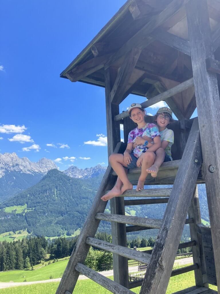 Two smiling children sitting in a wooden lookout structure with a bright blue sky and a picturesque alpine landscape in the background. The scene features lush green hills, tall pine trees, and jagged mountain peaks under a sunny summer day.