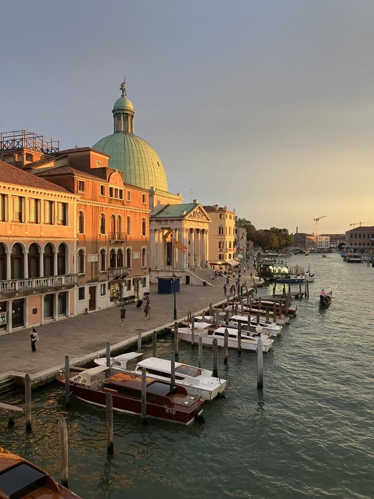 A scenic view of a canal in Venice during sunset, featuring colorful buildings on the left with a green-domed church. Small boats are docked along the waterfront, while a few boats navigate the canal. The sky is a blend of warm hues.