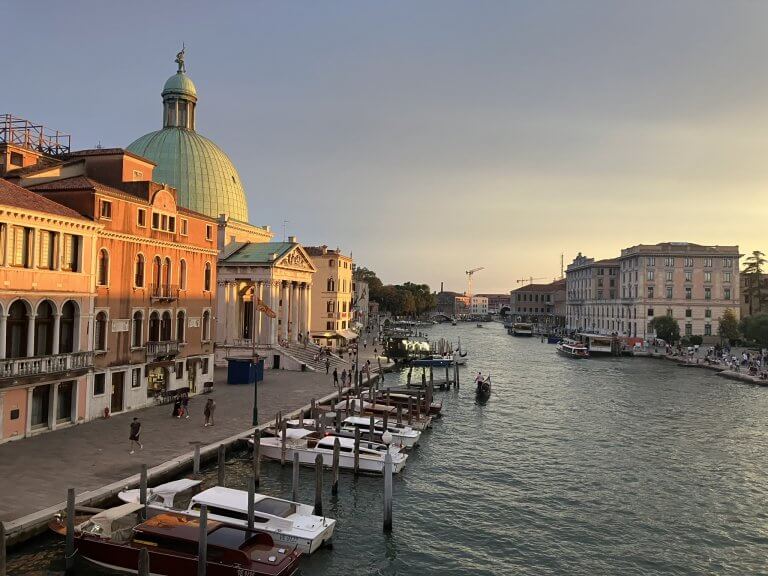 A scenic view of a canal in Venice during sunset, featuring colorful buildings on the left with a green-domed church. Small boats are docked along the waterfront, while a few boats navigate the canal. The sky is a blend of warm hues.