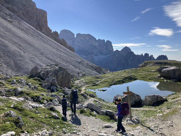 Two hikers walk along a rocky trail near a small reflective pond surrounded by grass and boulders, with jagged mountain peaks rising dramatically in the distance. A wooden trail sign stands by the path, and the bright blue sky enhances the scenic alpine setting.
