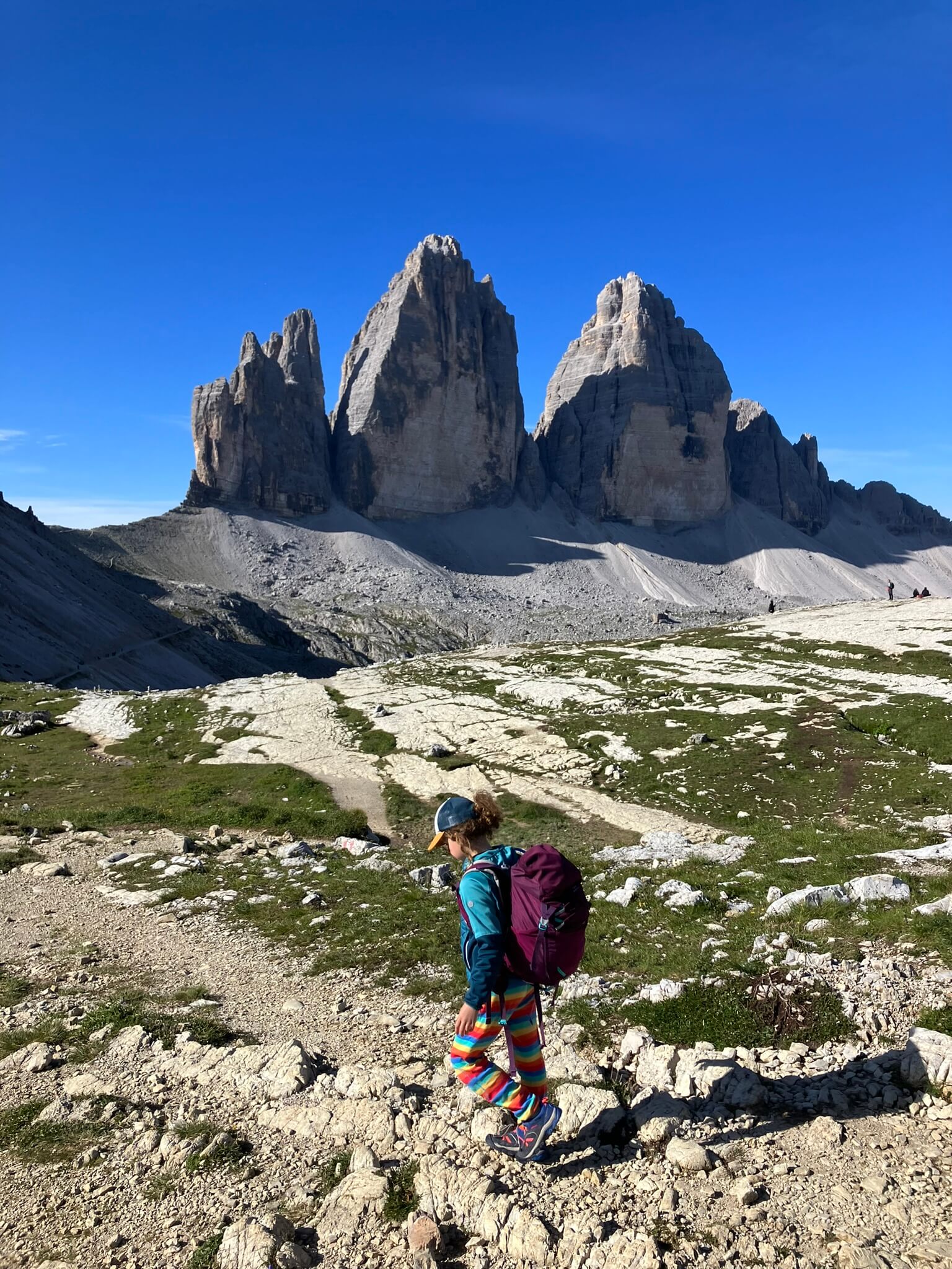 A child wearing colorful striped pants and a backpack trekking through a rugged landscape in the Dolomites, with the iconic Tre Cime di Lavaredo rising dramatically against the clear blue sky. This captures the unique beauty and excitement of a hut to hut hike with kids in the Dolomites.