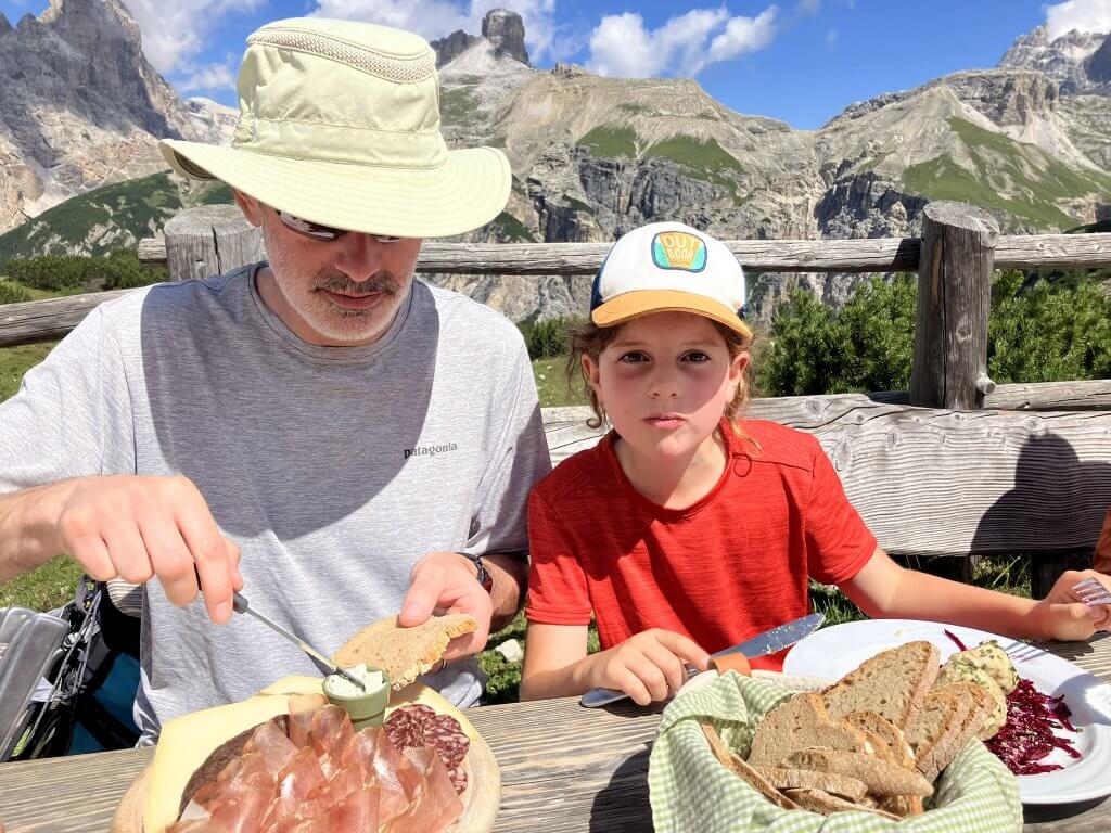 A man and child enjoying a hearty meal outdoors at a wooden picnic table during a hut to hut hike in the Dolomites. The plate features local delicacies like cured meats, cheese, and bread, with majestic mountain scenery in the background.