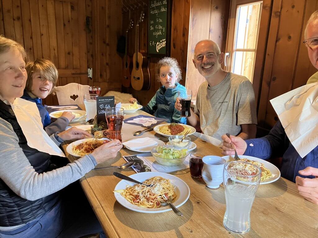 A group of six people sitting at a wooden table in a cozy room, enjoying plates of pasta. Two guitars hang on the wood-paneled wall behind them. They are smiling and have drinks in front of them, including a glass of lemonade.