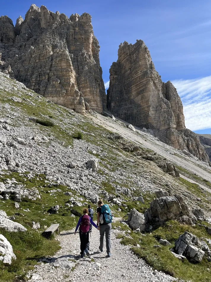 Two hikers walk along a rocky trail surrounded by grassy patches and scattered stones, leading toward towering jagged rock formations under a bright blue sky. The hikers wear backpacks, with one appearing to be an adult and the other a child, emphasizing a shared outdoor adventure in a dramatic alpine landscape.