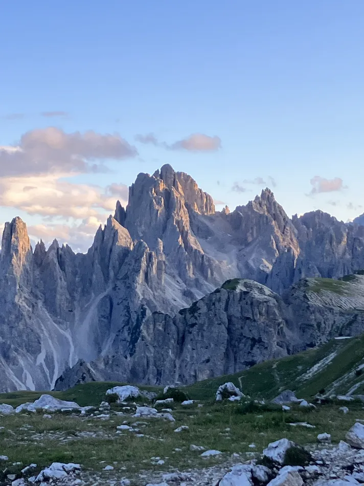 A stunning view of jagged mountain peaks bathed in warm, golden light against a clear blue sky. The foreground features patches of green grass and scattered rocks, adding to the rugged charm of the alpine scenery.
