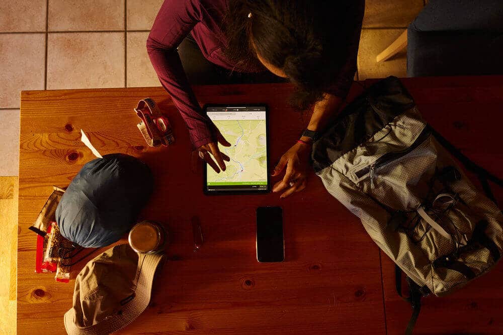 A person plans a hiking route on a tablet at a wooden table filled with outdoor gear, including a backpack, hat, snacks, and a water bottle. The setting suggests preparation for an adventure.