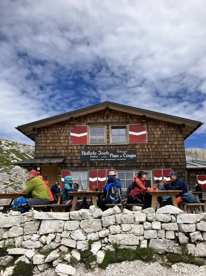 A mountain hut with a wooden exterior and red and white shutters sits under a cloudy sky. People in outdoor gear are seated at picnic tables in front, enjoying the scenic setting. Stone walls and alpine terrain surround the area.