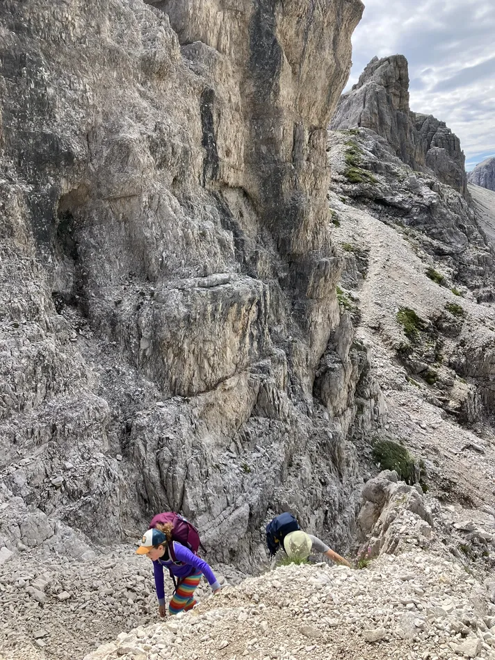 Two people are climbing a steep rocky terrain, wearing backpacks. One wears a colorful striped outfit and cap, while the other is dressed in neutral tones. The background shows rugged rock formations. The scene suggests a challenging outdoor hike.