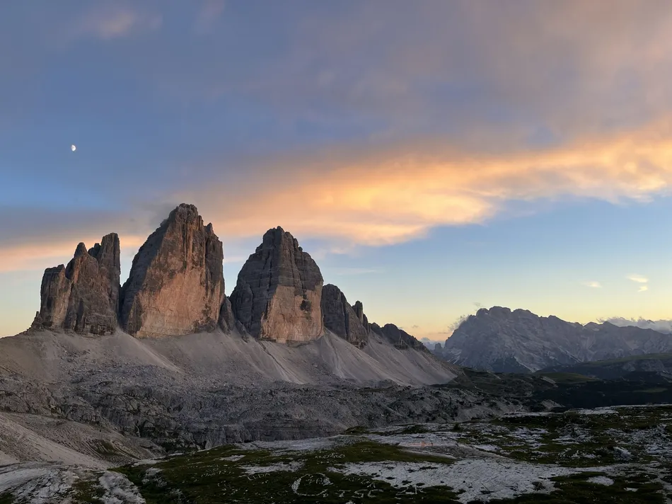 A stunning sunset view of the Tre Cime di Lavaredo peaks in the Dolomites, with the moon visible in the sky and soft orange and pink clouds stretching across the horizon. The rugged terrain and grassy patches create a dramatic backdrop for a hut-to-hut hike in the Dolomites with kids.