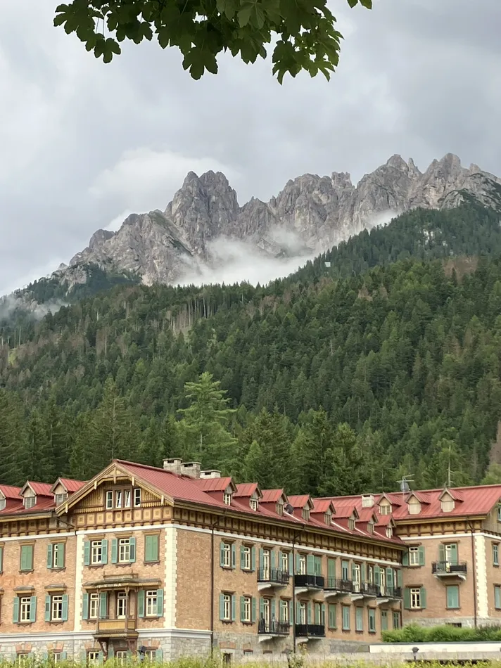 A large, grand building with a red roof set against a backdrop of lush green forest and jagged mountain peaks under a cloudy sky. A large tree frames the top of the image.