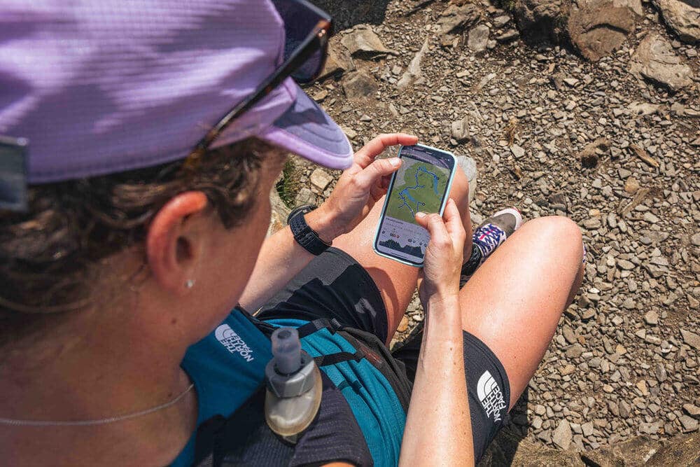 A person wearing a purple cap and black athletic gear is sitting on rocky terrain while looking at a Komoot map on a smartphone. They have a hydration pack on their back and colourful sneakers.