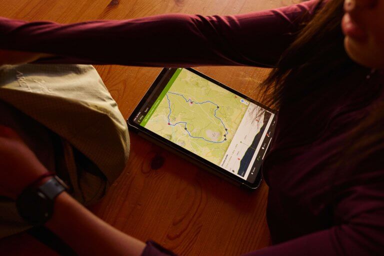 A person in a maroon top is sitting at a wooden table, looking at a tablet showing a hiking route on a map. Their arms are partially visible, and they have a watch on their wrist. There is a backpack nearby.
