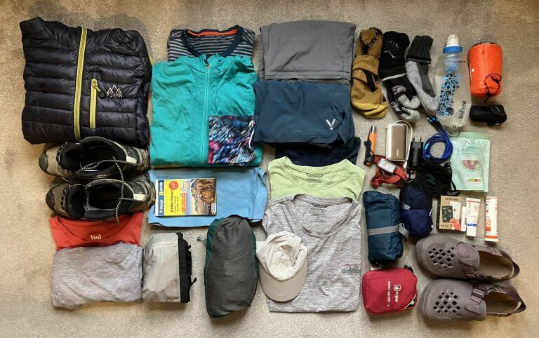 A neatly organized flatlay of a complete packing list for hut hiking, featuring clothing layers, hiking boots, a water bottle, sunscreen, snacks, and essentials like a first-aid kit and map.