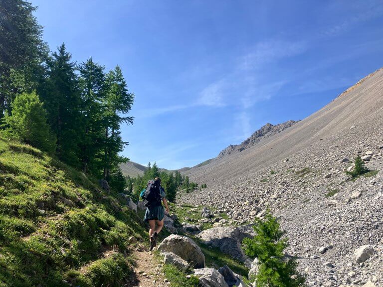 A hiker walks along a dirt path surrounded by a mix of grassy slopes and rocky terrain, with a bright blue sky overhead and alpine peaks in the distance, a scenic moment on a 3-day hiking itinerary in France.