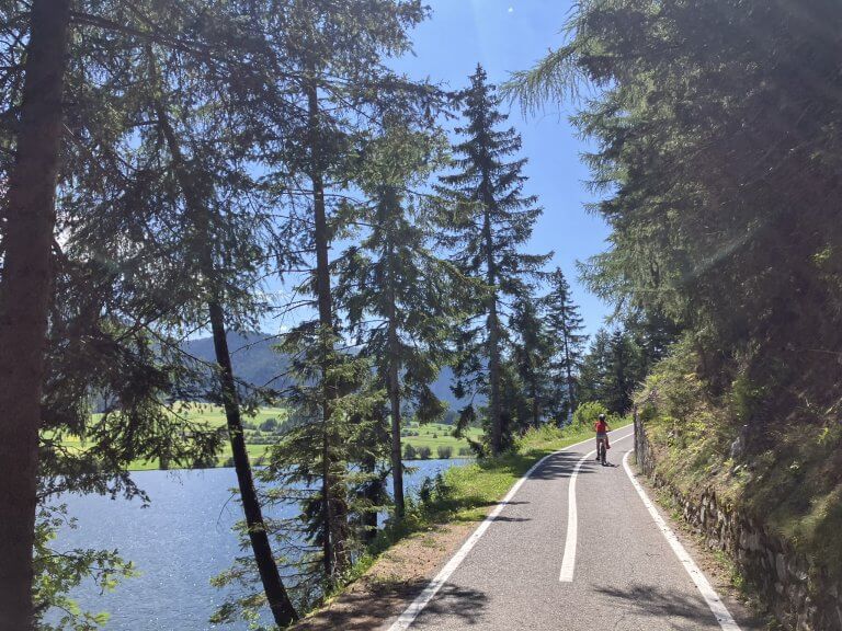 A cyclist pedalling along the Adige Cycle Path shaded by tall trees, with a sparkling lake visible through the branches. The natural beauty emphasizes why cycling the Adige Cycle Path with kids is an unforgettable experience.