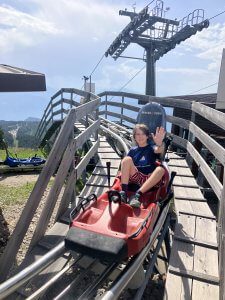 A child waves from a red alpine coaster, with scenic mountain views in the background. One of the 15 best things to do with kids in Lienz.