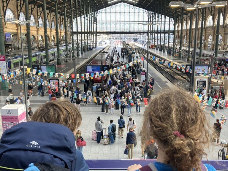 A view from the upper level of a grand train station with travelers and trains below. The large arched windows and glass-paneled roof let in natural light as two children with backpacks look over the railing.
