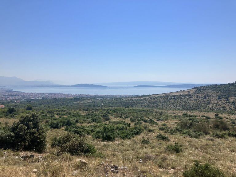 A high-altitude view of a coastal city with the sea stretching into the horizon. The landscape includes patches of green, dry land, and buildings clustered near the water’s edge.