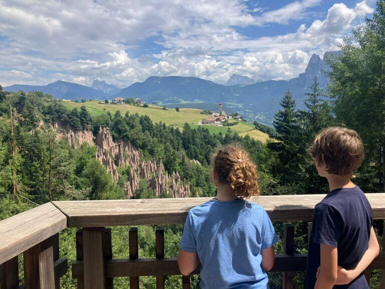 Two children stand at a wooden viewpoint railing, gazing at the Earth Pyramids of Ritten, with lush green hills, a small church, and the Dolomites in the background.