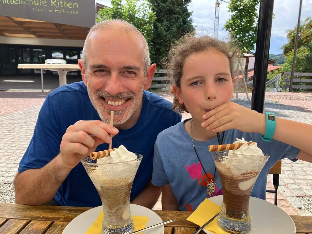 A father and daughter smile while sipping iced coffee topped with whipped cream and wafer rolls at an outdoor café in Ritten, Italy.