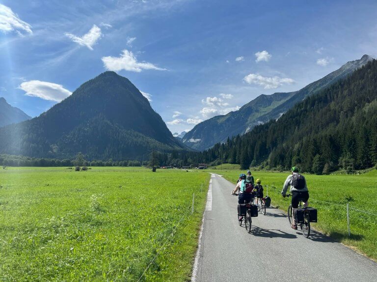 Cyclists enjoying a bike holiday in Austria, riding along a picturesque alpine valley with green meadows and majestic mountains under a clear blue sky.