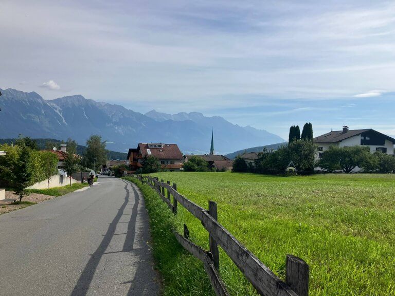 A scenic countryside road bordered by a wooden fence, leading to the village of Mutters - the best rural place to stay for access to Innsbruck - with houses and a church spire, set against a backdrop of majestic mountains under a partly cloudy sky.