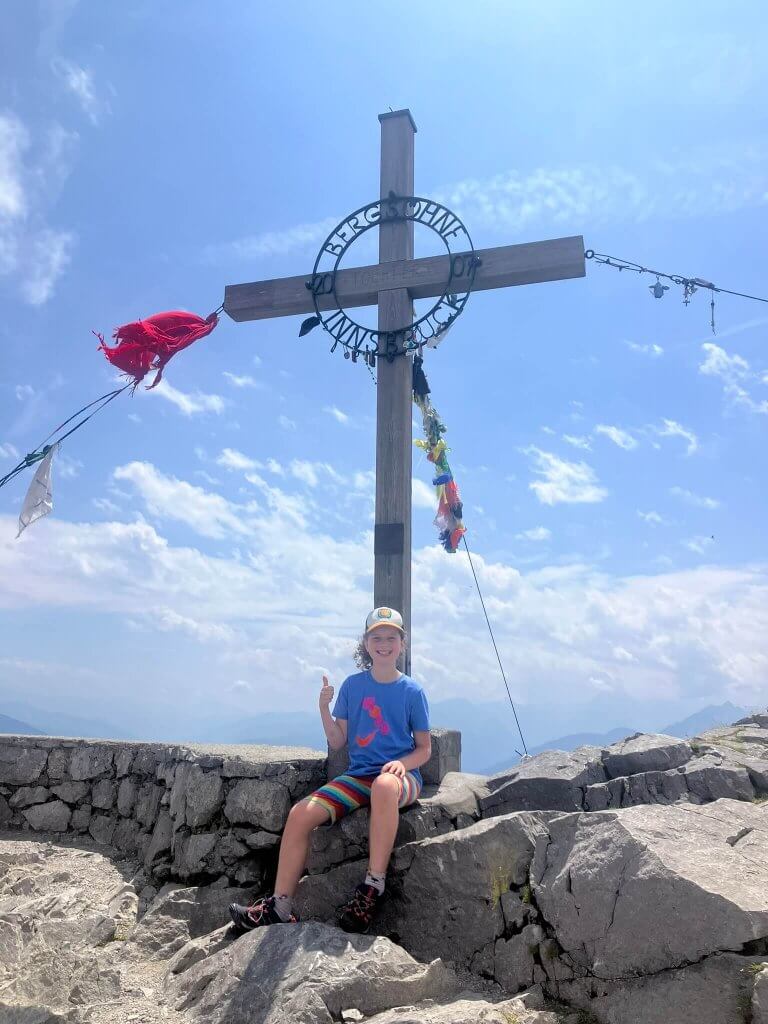 A child sitting on a rocky summit near Innsbruck, next to a large wooden cross with panoramic views of the Alps—a great day out from Innsbruck.