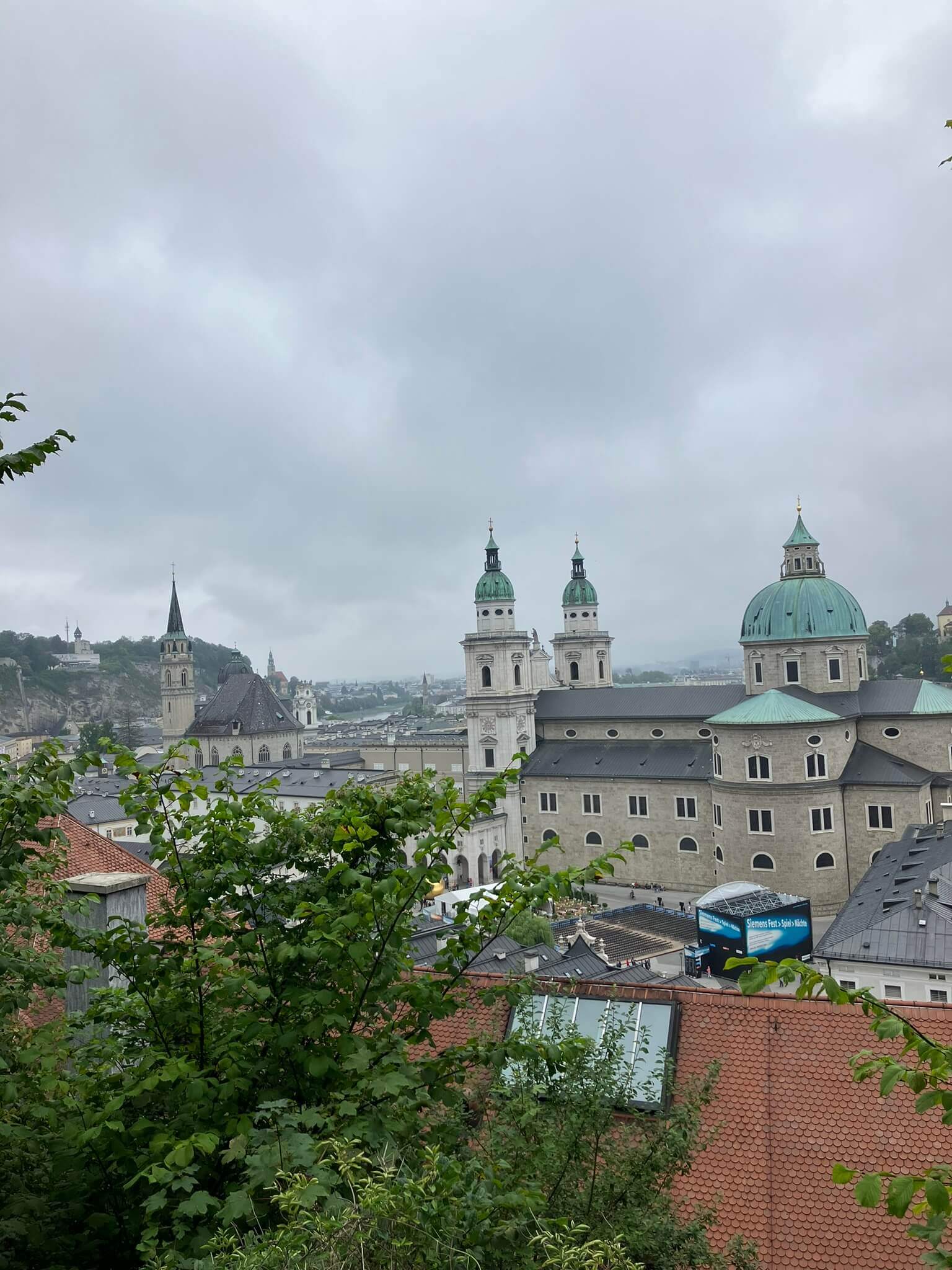 Panoramic view of Salzburg's old town, with the cathedral's green domes and surrounding historic buildings framed by cloudy skies and greenery in the foreground.