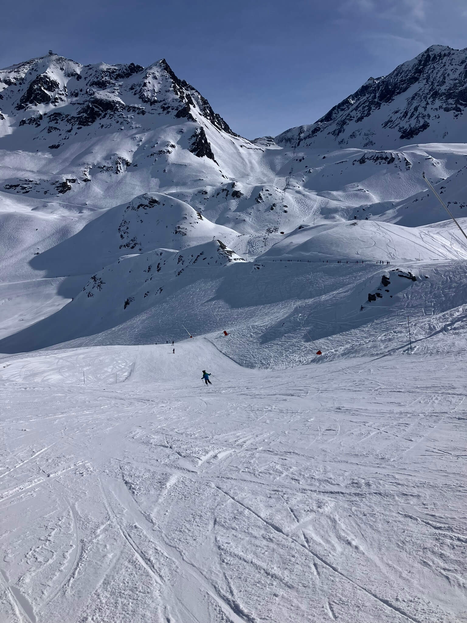 A vast snow-covered mountain landscape with a skier in the distance. The sky is blue and the sun is shining on a glorious bluebird day in the Easter holidays.