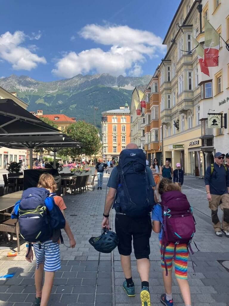 A family walking through the streets of Innsbruck with hiking backpacks, surrounded by colorful buildings and mountain views.