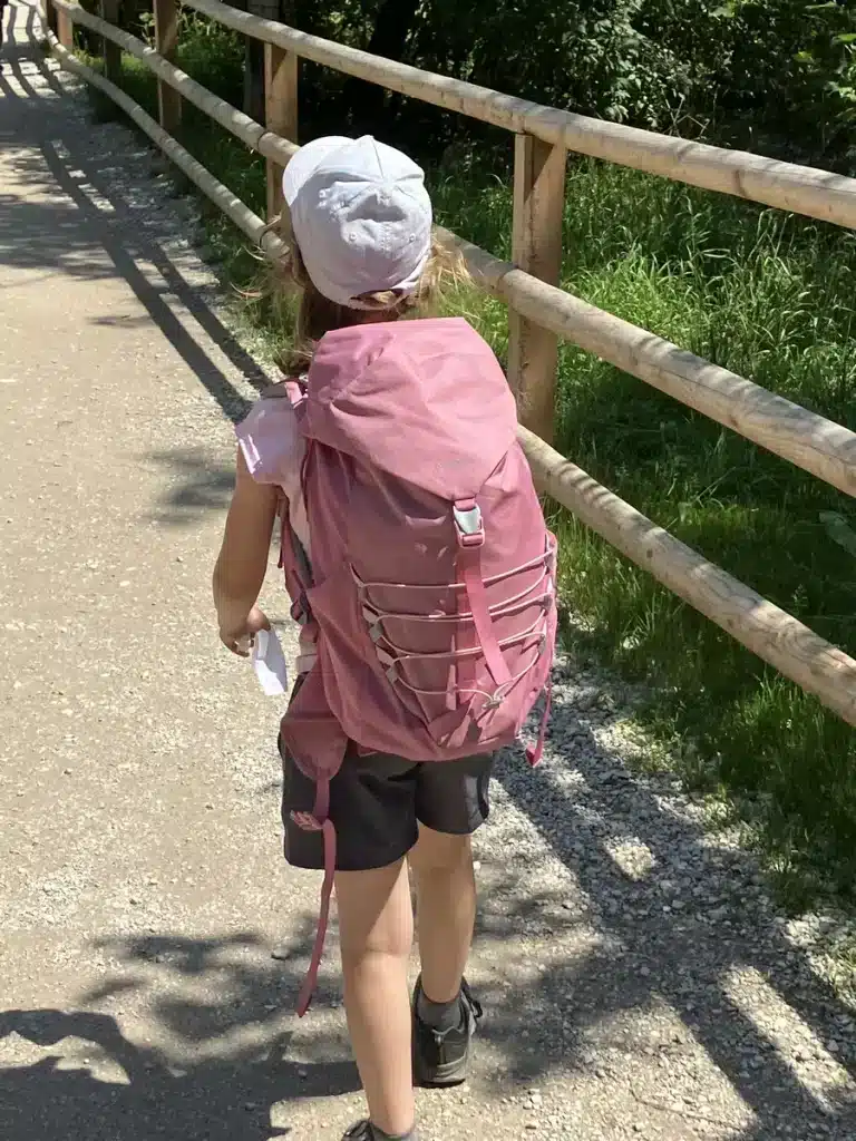 A young girl in a white cap and pink shirt carries a bright pink hiking backpack while walking on a shaded gravel path. A practical look at the best hiking backpacks for kids on the trail.