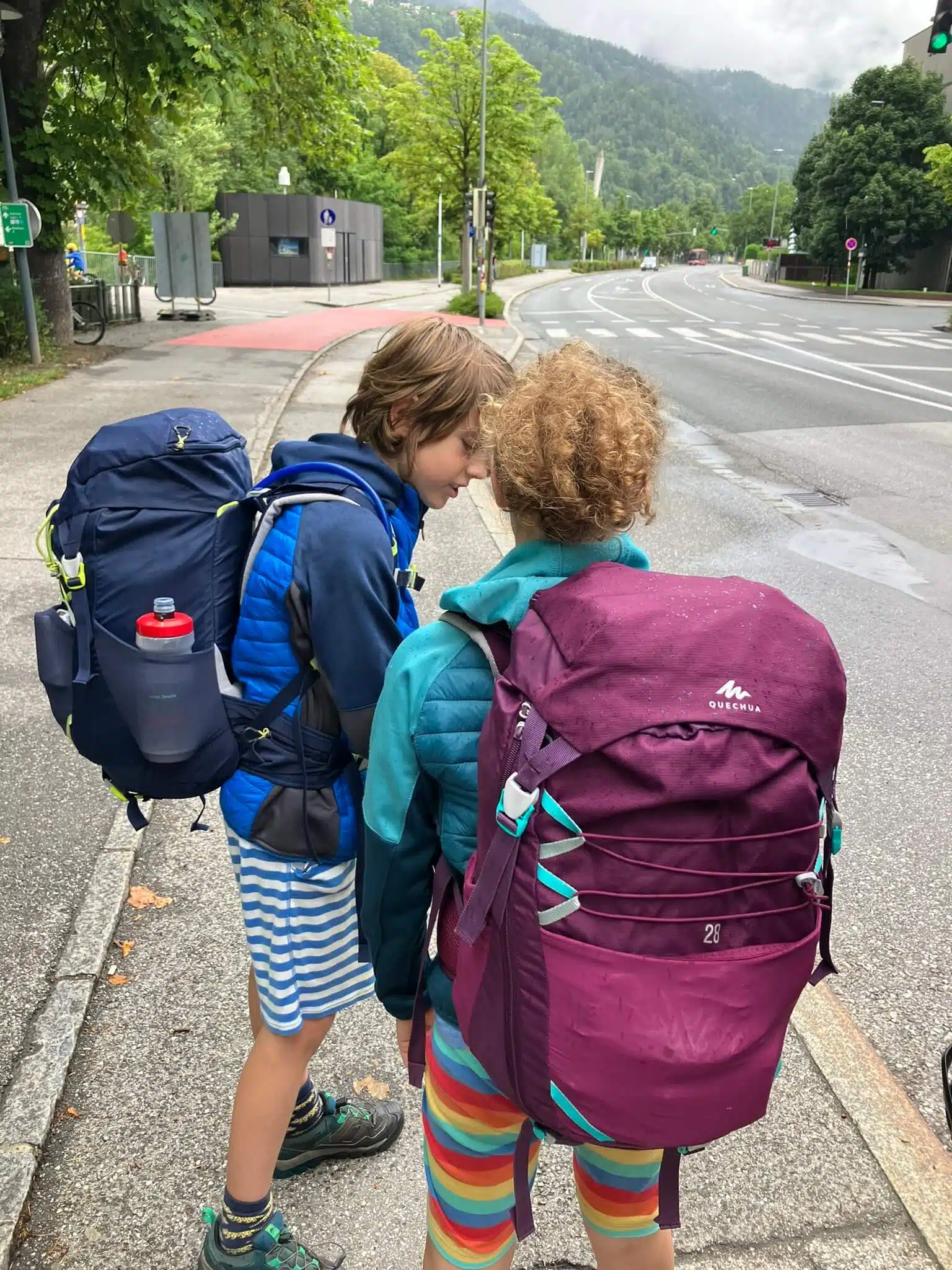 Two children wearing colorful outfits and carrying Quechua hiking backpacks, including a maroon 28L model, stand on a sidewalk with a scenic mountain backdrop, demonstrating real-world use of some of the best hiking backpacks for kids.