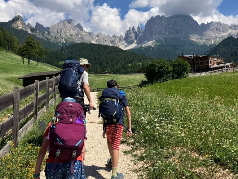 A family hikes along a gravel trail in the Dolomites under a bright blue sky, with two children carrying large, well-fitted hiking backpacks, highlighting some of the best hiking backpacks for kids. Majestic rocky peaks and a cozy mountain lodge are visible in the scenic background.