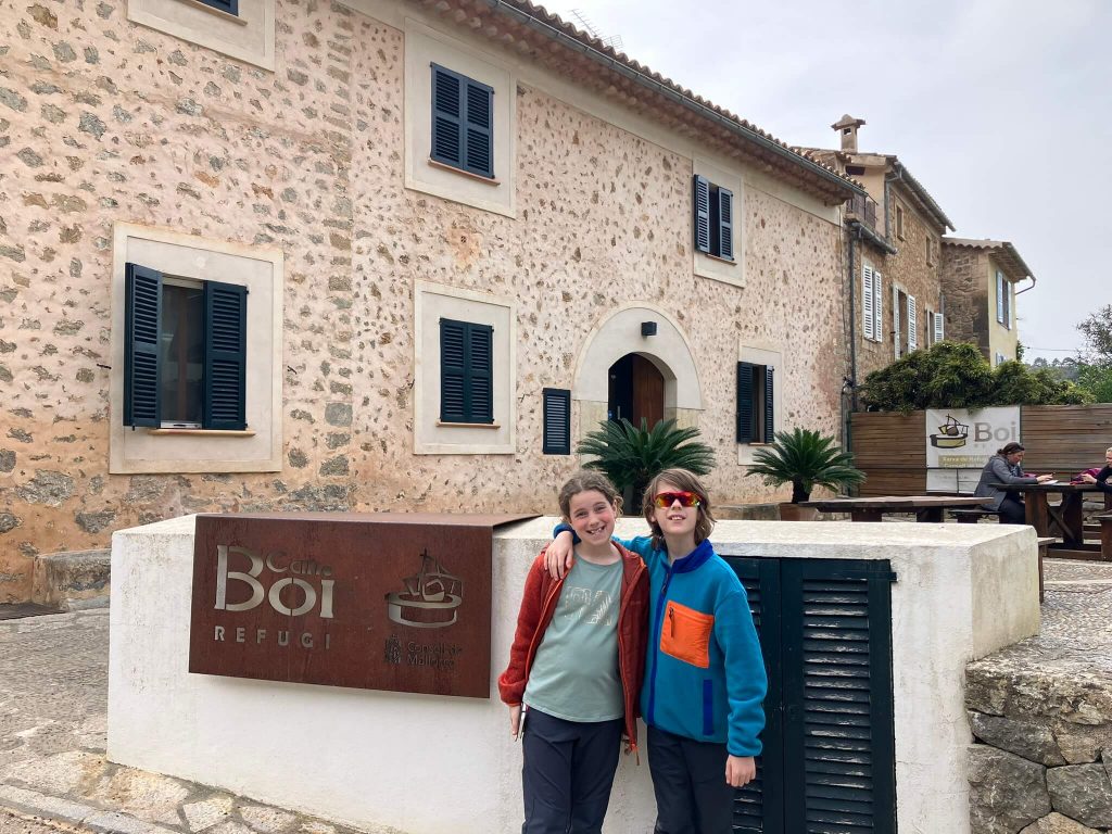 Two kids smile in front of the Can Boi refuge in Deià, a charming stone building with blue shutters—one of the most family-friendly stops on the GR221 Mallorca hiking itinerary for beginners or families.