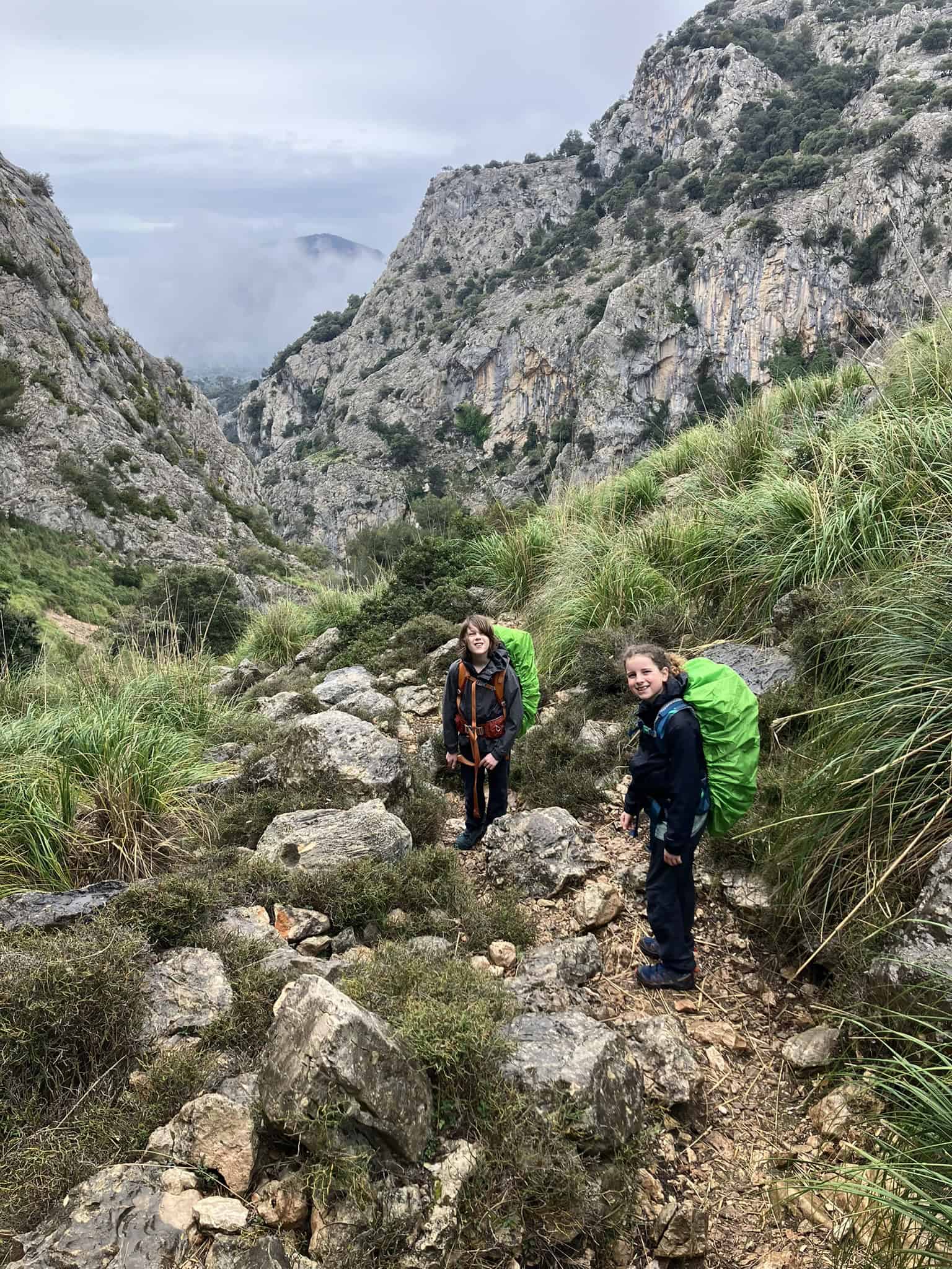 Two kids navigate a rugged, misty mountain pass surrounded by dramatic cliffs and wild greenery—challenging but rewarding terrain off the GR221’s main route.