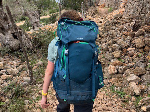 The back view of a young hiker navigating a rocky path, showing off the full frame and loaded structure of the Quechua 40+10L Kids Hiking Backpack in motion.