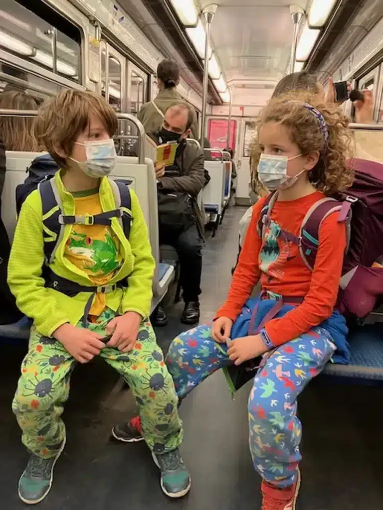Two children wearing face masks and backpacks ride a busy Paris metro, an everyday moment in raising resilient children through family travel and adapting to new environments.