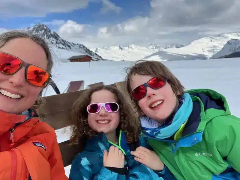 A snowy mountain backdrop frames a smiling mum and two kids in bright ski gear and colorful sunglasses, seated on a bench during a ski day. Embracing Easter skiing while travelling adventurously as a solo parent with kids.