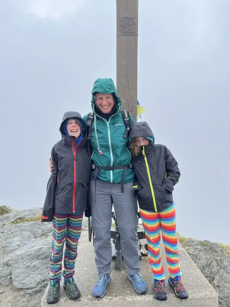 A mom and two kids stand at a foggy mountaintop, smiling despite the rain—a perfect example of raising resilient children through family travel and shared perseverance.