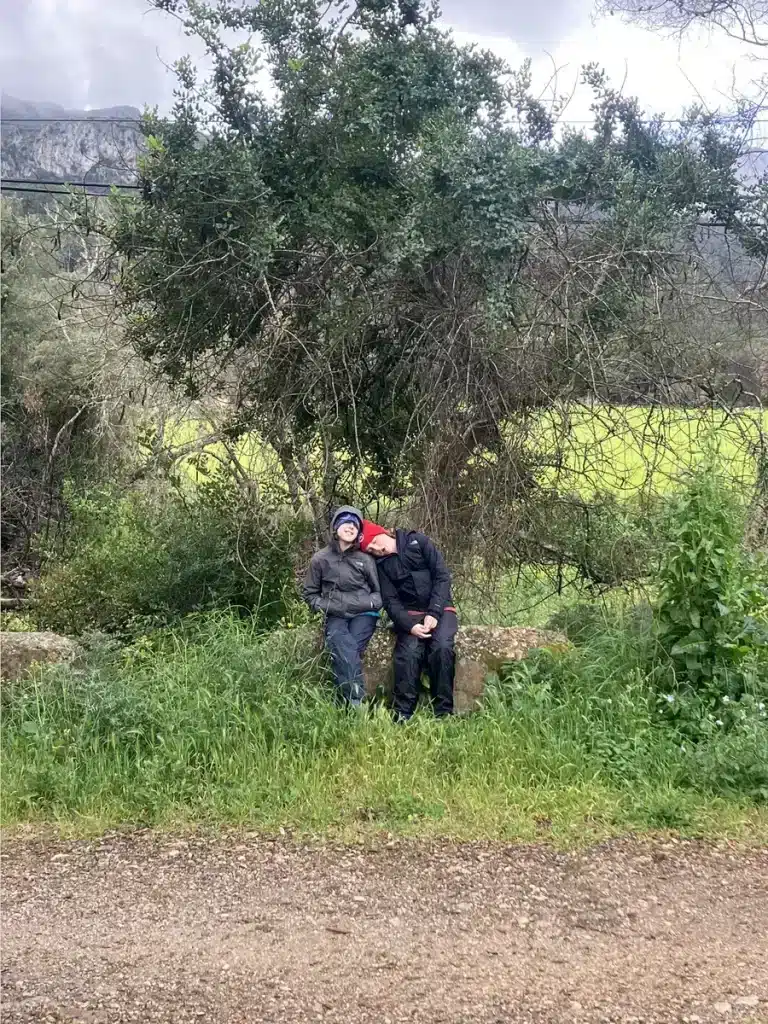 Two family members huddle together on a mossy bench, waiting in the rain—an intimate scene showing how raising resilient children through family travel means finding comfort anywhere.